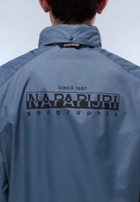 Lichtblauwe waterdichte jas met een hoge kraag, zwart logo tekst "NAPAPIJRI" en "SINCE 1987" prominent gedrukt op de achterkant.