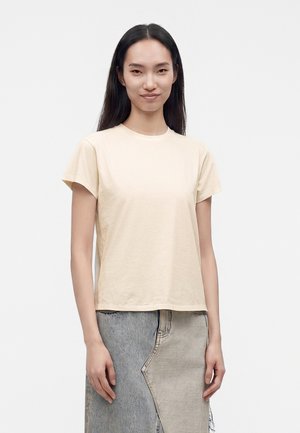 Jeune femme aux longs cheveux noirs portant un t-shirt beige uni à manches courtes et une jupe en jean bicolore, debout devant un fond blanc.