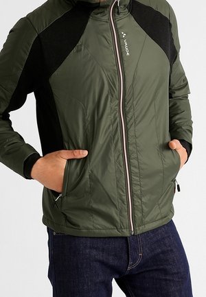 Homme portant une veste zippée verte et noire, les mains dans les poches avant, et un jean bleu foncé, sur un fond uni.