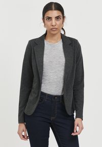 Tmavě šedý blazer vyrobený z měkké tkaniny, s jedním knoflíkem, s klopami, zkombinovaný se světle šedým tričkem a tmavými džíny.