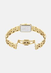 Zlata metalna ura s pravokotnim ekranom, povezano verižico, vidnim zapiranjem in logotipom na hrbtni strani. Minimalističen dizajn, poliširan zaključek.