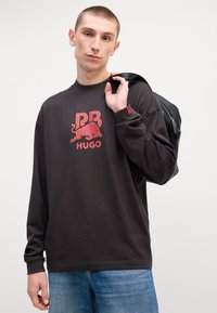 T-shirt scura a maniche lunghe con una grafica rossa di un logo di toro con il testo "HUGO", abbinata a jeans in denim blu chiaro e una borsa nera.
