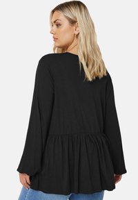 Top nero a maniche lunghe con design a peplum a strati, tessuto morbido, scollatura a barca e vestibilità comoda. Abbinato a jeans azzurri.