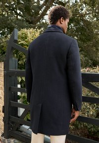 Cappotto di lana scuro con vestibilità sartoriale, caratterizzato da una sola apertura, revers a dente e polsini con bottoni, mostrato da dietro in un contesto di giardino.