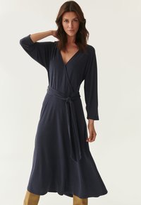 Navy blue wrap dress in pehmeä kangas, jossa on V-aukko, pitkät hihat ja solmittava vyötäröyksityiskohta. Liikkuva hame ulottuu puoleen pohkeeseen.