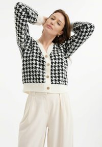 Svetr s vzorem houndstooth v černobílé barvě, s výstřihem do V, krémovými knoflíky a žebrovanými manžetami, v kombinaci se světle béžovými kalhotami.