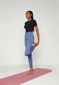 Camiseta preta ajustada e leggings azul claro, de pé descalça sobre um tapete de yoga rosa, a realizar uma pose de equilíbrio. Fundo neutro.