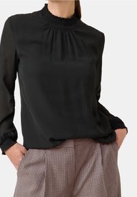 Schwarze Bluse mit hochgezogenem, gerafftem Kragen, langen Ärmeln mit gerafften Manschetten, kombiniert mit karierter Hose. Leicht, weiche Textur.