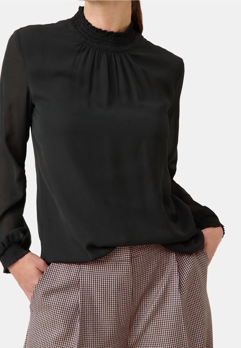 Schwarze Bluse mit hochgezogenem, gerafftem Kragen, langen Ärmeln mit gerafften Manschetten, kombiniert mit karierter Hose. Leicht, weiche Textur.
