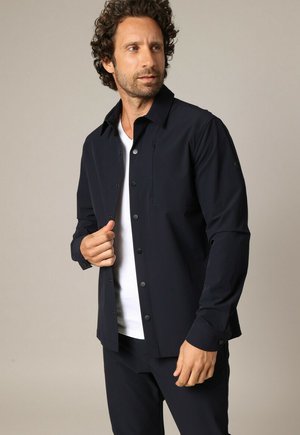 Man met krullend haar die een zwart overhemd met knopen draagt over een wit T-shirt, het overhemd met één hand verstelend, en zijwaarts kijkend.