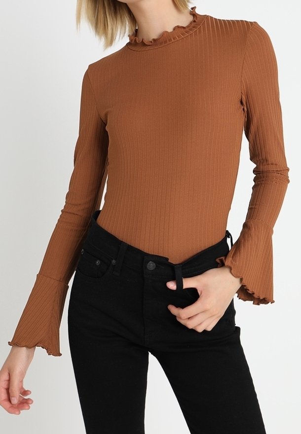 Femme portant un haut ajusté en rib court marron à manches longues avec des poignets et un col volantés, associé à un jeans noir taille haute.
