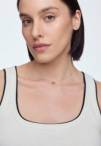 Złoty naszyjnik z okrągłym wisiorkiem, noszony na jasnym topie na ramiączkach z ciemnymi wykończeniami. Gładka tekstura i minimalistyczny design.