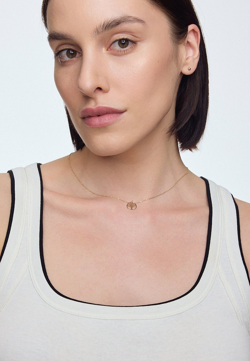 Złoty naszyjnik z okrągłym wisiorkiem, noszony na jasnym topie na ramiączkach z ciemnymi wykończeniami. Gładka tekstura i minimalistyczny design.