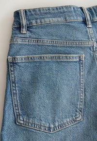 Poche arrière et ceinture d'un jean denim bleu avec coutures visibles et passants de ceinture sur un fond clair.