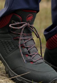 Scarpa da trekking di un verde scuro con accenti rossi, tomaia texturizzata e lacci pattern. Presenta una suola robusta e un tallone rinforzato per supporto.