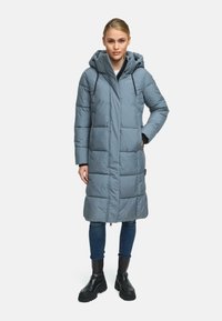 Manteau d'hiver long gris avec capuche, présentant des panneaux matelassés horizontaux et des accents en métal noir. Porté sur un jean et des bottes noires.
