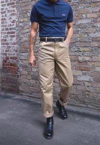 Marinblå polo skjorta, khakisbyxor, svart bälte och ankelstövlar. Tegelvägg i bakgrunden. Byxorna är uppvikta vid fållen, vilket ger en skräddarsydd look.