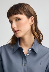 Camicia col polo di colore azzurro chiaro in tessuto liscio con piccoli bottoni bianchi, dotata di un logo ricamato sul lato sinistro.