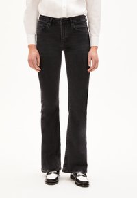 Black denim flared jeans with a mid-rise waistband, five pockets, and a soft texture. Paired with black and white shoes.