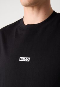 T-shirt in cotone nero con collo a giro. Presenta una patch rettangolare bianca con la scritta "HUGO" sul petto. Texture morbida, vestibilità casual.