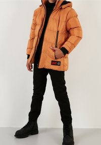 Chaqueta acolchada naranja con capucha, cremallera frontal y detalles contrastantes en negro. Se lleva sobre una camiseta negra, combinada con pantalones y botas negras.