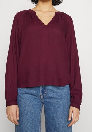 Frau trägt eine lange, locker sitzende violette Bluse mit V-Ausschnitt und lange Ärmel, kombiniert mit blauen Jeans, dargestellt von Hals bis Hüfte vor einem schlichten Hintergrund.