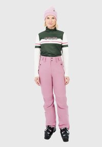 Vrouw in roze skibroek, zwarte skischoenen, groene en witte lange mouwen top met de tekst "SKI YA LATER" en een roze beanie.