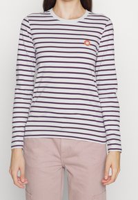 Långärmad randig skjorta i vitt och lila, gjord av bomull. Har en rund halsringning och ett litet broderat märke på bröstet.