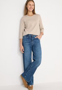 Chemise beige à manches longues associée à un jean denim bleu à jambes larges, avec une poche avant et une coupe décontractée. Des baskets marron complètent la tenue.