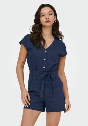 Frau mit langen dunklen Haaren, die einen marineblauen kurzärmeligen Overall mit Knöpfen und Taillengürtel trägt, steht mit einer Hand nahe der Hüfte.