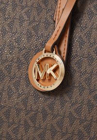 Sac à main en cuir marron avec un design texturé, quincaillerie dorée et un logo circulaire en relief avec les initiales "MK" et le texte "Michael Kors".