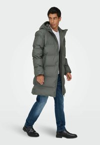 Manteau long rembourré vert foncé avec une capuche, doté d'une fermeture éclair, de poches latérales et d'une coupe décontractée, porté sur une chemise blanche et un jean.