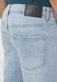 Φωτεινά μπλε τζιν φορεμένα από ένα άτομο, με εμφανή την πίσω τσέπη και μαύρο δερμάτινο ετικέτα στην μέση.