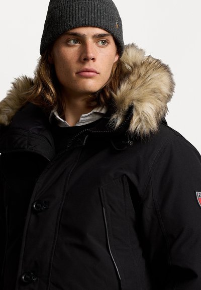 Črna parka jakna s kapuco obloženo s krznom, ki ima gumbasto zapiranje, stranske žepe in logotip obliž. V kompletu s sivo rebrasto kapa.