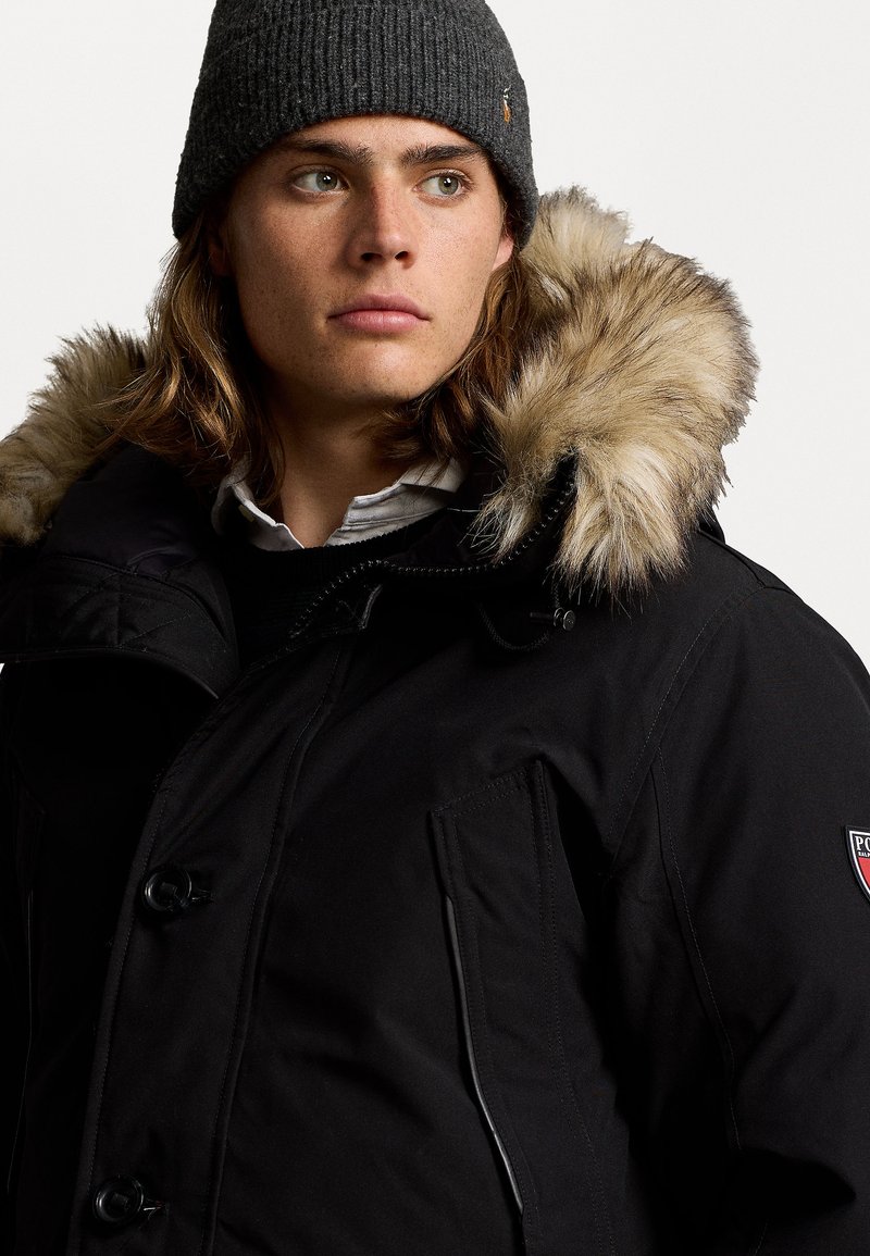 Parka nera con cappuccio foderato in pelliccia, dotata di chiusura con bottoni, tasche laterali e una patch con logo. Accompagnata da un berretto grigio a coste.