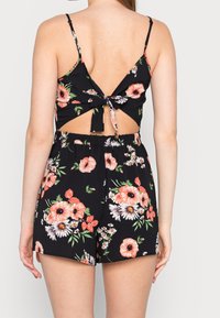 Kwiatowy kombinezon w kolorze czarnym z otwartym plecami, z detalem w postaci wstążki oraz elastyczną talią. Zawiera duże różowe, białe i czerwone kwiaty z zielonymi liśćmi.