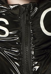 Svart puffjacka med glänsande finish, framsida med dragkedja och märkesdragare, samt stor vit text på tyget.