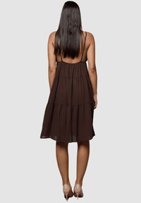 Vestido marrón con capas y tirantes finos, hecho de un tejido ligero. Presenta un corte holgado y llega hasta la mitad de la pantorrilla, combinado con tacones nude.