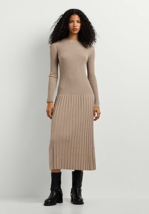 Donna con capelli ricci che indossa un lungo vestito maglione beige a coste e stivali neri a metà polpaccio, in piedi contro una parete chiara e semplice.