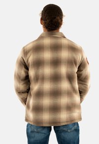 Chaqueta de cuadros marrón con una tela texturizada, que presenta un collar y un parche con logo en la parte posterior. Combinada con jeans azules, mostrando un corte relajado.