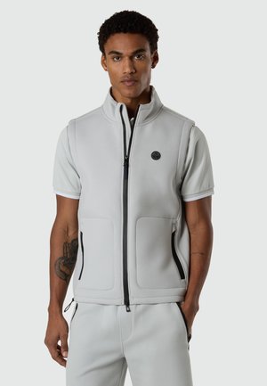 Man wearing light gray sleeveless zip-up vest over matching short-sleeve shirt and pants, standing with a neutral expression.