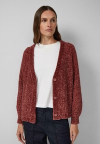 Texturierter, ziegelroter Cardigan mit geripptem Design, offenem Frontverschluss und vier großen Knöpfen. Darüber getragen ein weißes Hemd und eine dunkle Hose.