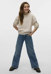 Pull beige avec des pois texturés, col rond et manches retroussées, associé à un pantalon bleu large et des chaussures noires. Fond simple.