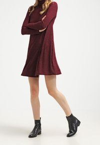 Robe en tricot côtelé bordeaux avec col cheminée, forme évasée et manches longues. Portée avec des bottes de cheville noires à talon bas.