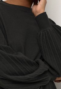 Eine Person trägt einen schwarzen Pullover mit weiten, gerippten, gefalteten Ballon�ärmeln und einem gerippten Halsauschnitt und berührt mit einer Hand ihr Gesicht.