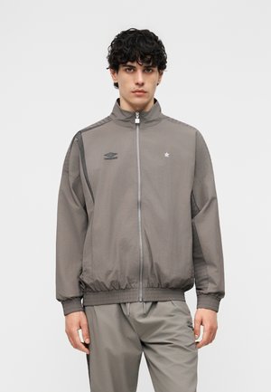 Ung mann iført grå Umbro treningsjakke og matchende bukser, stående foran en ensfarget lys bakgrunn.