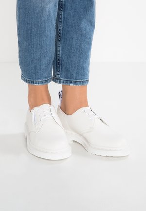 Hvide teksturerede lædersko med snørebånd og lynlåse i siden, båret med blå korte jeans på en hvid gulv.