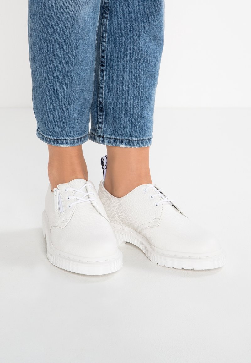 Chaussures blanches en cuir texturé à lacets avec fermetures éclair latérales portées avec un jean bleu court sur un sol blanc.