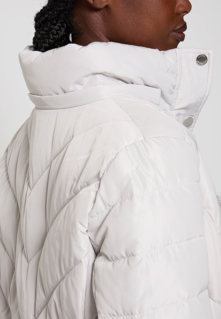 Biała pikowana kurtka z wysokim kołnierzem, wykończona napami. Gładki, błyszczący materiał o miękkiej teksturze.