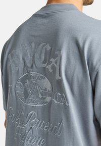 Grijze katoenen T-shirt met geborduurde tekst en een wereldbolafbeelding op de achterkant. Het ontwerp bevat een motief van "Verleden Heden Toekomst".